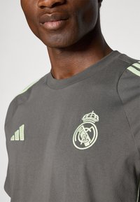 Grå kortærmet t-shirt med lysegrønne Adidas striber og Real Madrid logo, med rund halsudskæring og blødt bomuldsstof.