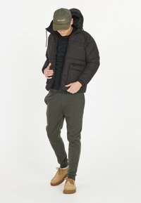 Man som bär en svart puffjacka, svart skjorta, olivgröna sweatpants, bruna skor och en olivgrön keps står framför en vit bakgrund.