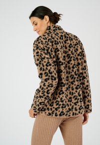 Leopardprint fleece jack met tan en zwarte vlekken, voorzien van een kraag en een gestructureerde stof, gecombineerd met ribbelige tan broeken.