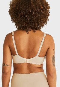 Vrouw met krullend haar in een beige bh en hoog uitgesneden ondergoed, met tatoeages op beide bovenarmen, tegen een effen achtergrond.