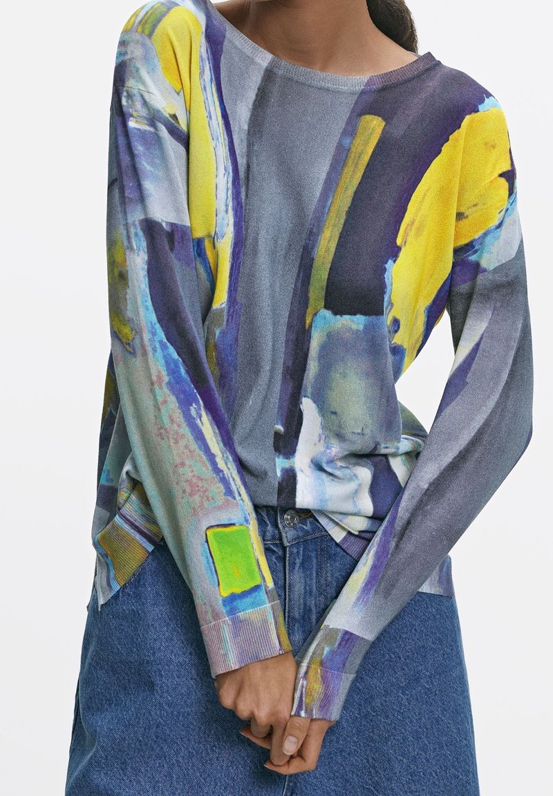 Femme portant un pull à manches longues avec des motifs abstraits jaunes, violets et bleus, associé à une jupe en denim bleu, mains jointes devant.