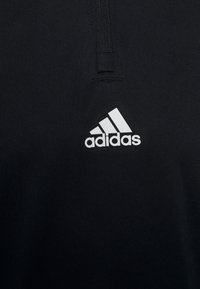 Zwarte sporttop met ritskraag, gemaakt van gladde stof, met een wit Adidas-logo aan de onderkant links.