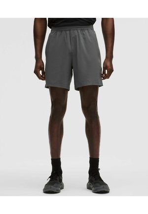 Mann trägt dunkelgraue Sportshorts, schwarze Socken und schwarze Laufschuhe und steht vor einem schlichten, hellen Hintergrund.