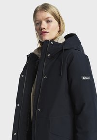 Femme blonde portant un manteau d'hiver à capuche navy foncé avec des poches avant et le logo AIGLE sur la manche, sur un fond clair et uni.