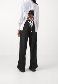 Bílá košile s dlouhým rukávem, otevřeným zadním dílem a zavazovacími detaily, kombinovaná s černými kalhotami s širokými nohavicemi, zdobenými bočními rozparky a texturovanou látkou.