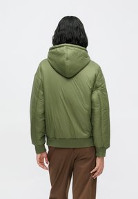 Podložena zelena jakna s kapuco, z ukrašeno obrobo in manšetami, gladka tekstura in sproščen kroj. Prikazan zadnji pogled.