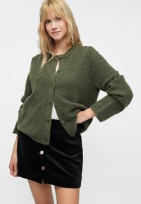 Ελιά πράσινο cardigan φτιαγμένο από μαλακό πλεκτό ύφασμα, με στρογγυλή λαιμόκοψη, κλείσιμο με κουμπί και χαμηλούς ώμους. Συνδυάζεται με μια μαύρη σουέτ φούστα.