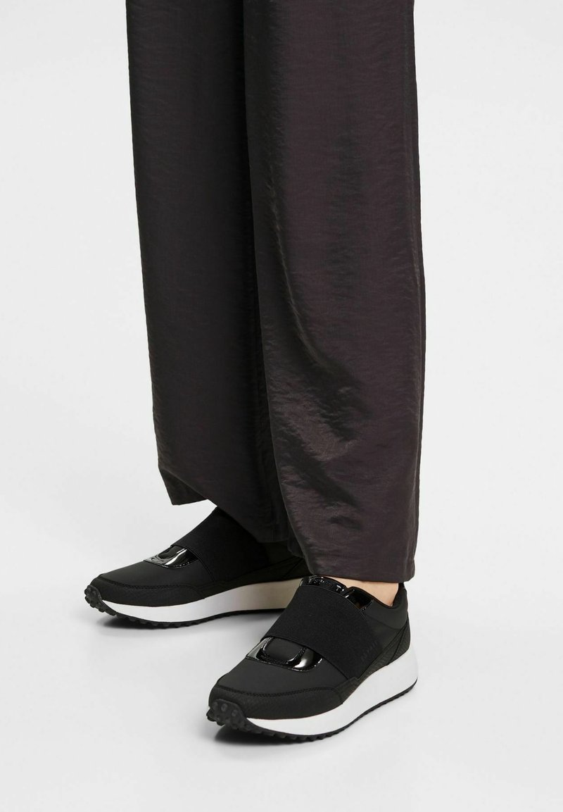 Baskets noires avec des sangles élastiques et des accents brillants, présentant un dessus texturé et des semelles blanches, accompagnées de pantalons larges sombres.