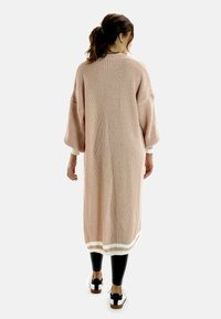Lång, rosa stickad kofta med lös passform, vida ärmar och vit kant i ärmsluten och nederkant, tillsammans med svarta leggings och sneakers.