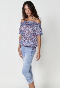 Blusa ampia a spalle scoperte con motivo floreale in blu, rosa e bianco; arricciatura elastica sul corpetto; indossata con pantaloni capri in denim chiaro.