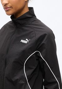 Γυναίκα που φοράει μαύρο αθλητικό μπουφάν Puma με λευκές ρίγες και φερμουάρ, ορατή από τους ώμους μέχρι το πηγούνι, μπροστά σε απλό φόντο.