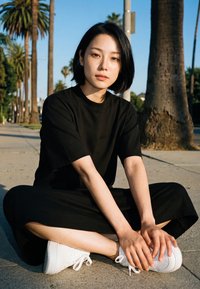 Ung kvinde med kort sort hår iført sort tøj, siddende med benene krydset på en solbeskinnet fortov, palmer og klar himmel i baggrunden.