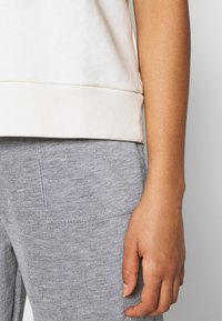 Sudadera de color beige claro con un dobladillo acanalado, combinada con pantalones de chándal grises de textura acanalada. Mano visible en el costado.