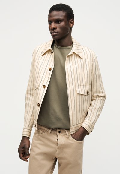 Homme portant une veste beige rayée boutonnée sur une chemise vert olive et un pantalon beige, debout avec une main dans la poche.