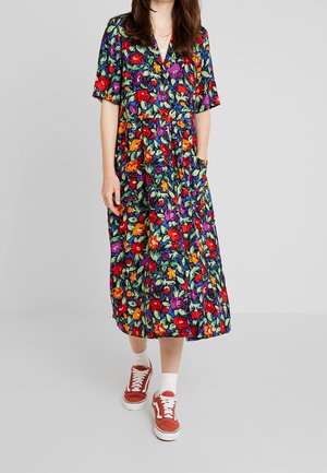 Femme portant une robe midi fleurie colorée à manches courtes, associée à des chaussettes blanches et des baskets rouges, debout devant un fond uni.
