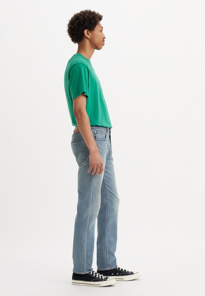 T-shirt in cotone verde, vestibilità rilassata, maniche corte. Jeans in denim blu chiaro, gamba dritta, finitura sbiadita. Sneakers in tela nera con suola bianca.