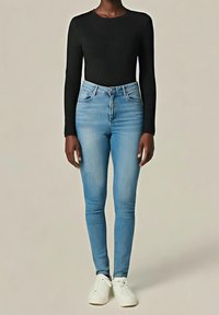 Femme portant un haut noir à manches longues, un jean skinny taille haute bleu clair et des baskets blanches, debout devant un fond neutre.
