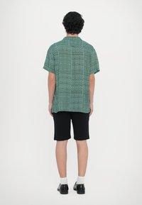 Camisa de manga curta verde com padrões geométricos, combinada com calções pretos. Modelo usa sapatos pretos e meias brancas.