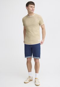 Tânăr stând și privind în lateral, îmbrăcat cu un tricou bej cu mânecă scurtă, pantaloni scurți din denim albastru închis, rulați, șosete albe și adidași bej.