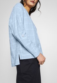 Blouse en lin bleu clair à manches longues, avec des rayures blanches, un col en V, des fentes latérales et un détail de bouton sur la manche.