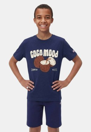 Ragazzo sorridente in piedi con le mani sui fianchi indossa un abbigliamento blu navy; la maglietta mostra noci di cocco e il testo "Coco Mood Champion Rules."