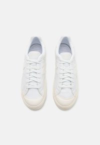 Baskets en cuir blanc avec une texture lisse, dotées d'une semelle en caoutchouc de couleur claire, de lacets plats et d'un design minimaliste avec un accent de logo.