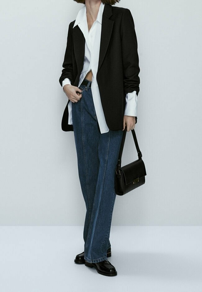 Blazer noir ajusté sur une chemise blanche, associé à un jean en denim noir à jambes larges et des chaussures en cuir noires. Petit sac à main noir avec une bandoulière.