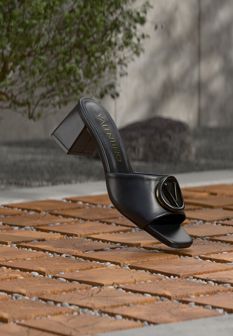 Μαύρο δερμάτινο σανδάλι με ανοιχτή μύτη και ψηλό τακούνι τύπου block, με χρυσό λογότυπο Valentino μπροστά, τοποθετημένο σε ξύλινη και πέτρινη εξωτερική επιφάνεια.