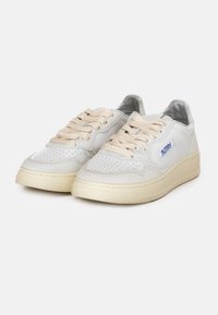 Sneaker in pelle bianca con punta perforata, lacci beige e dettagli metallici argento. Presentano una suola rialzata e un logo blu di lato.