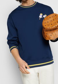 Sweat-shirt navy avec des accents jaunes au col et aux poignets, présentant deux designs de glace. Tenant un chapeau de seau beige avec des logos en relief.