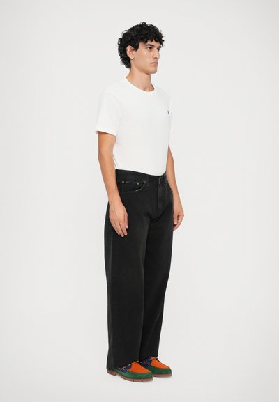 Camiseta branca, jeans largos pretos e sapatos de camurça multicoloridos com detalhes em laranja, verde e azul. Design simples e corte descontraído.