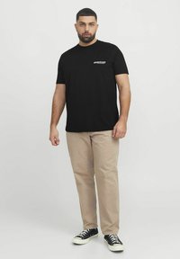 Camiseta de algodón negra de manga corta, con un pequeño logo blanco en el lado izquierdo del pecho. Combinada con pantalones beige y zapatillas negras.