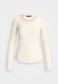 Długo rękawowa, kremowa bluzka z prążkowanego materiału ma okrągły dekolt oraz zapięcie na guziki z trzema beżowymi guzikami, wykończona szwem.
