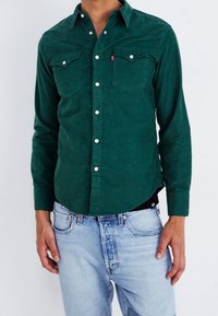 Camicia verde in velluto a coste con design a bottoni, due tasche sul petto e bottoni argentati, abbinata a jeans denim azzurri.