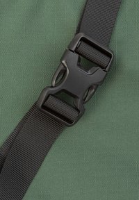 Nahaufnahme einer schwarzen Kunststoffschnalle auf einem schwarzen Riemen, vor einem grünen strukturierten Stoffhintergrund.