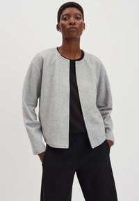 Lichtgrijze wollen blazer met een ronde halslijn, raglanmouwen en zonder sluitingen. Gecombineerd met een zwarte top en zwarte broek.