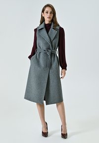 Gilet in misto lana grigia con vita cintata, ampie rever e tasche laterali. Indossato sopra una blusa bordeaux, abbinato a scarpe col tacco marroni.