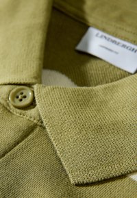 Polo en maille vert olive avec col boutonné, tissu texturé et une étiquette partiellement visible près du col.
