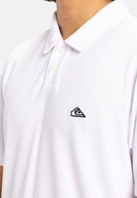Weißes Poloshirt aus strukturiertem Stoff, ausgestattet mit einem klassischen Kragen, zwei Knöpfen und einem schwarzen gestickten Logo auf der Brust.