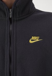 Svart fleecejacka med full dragkedja och gul Nike-logotyp broderad på vänster bröst.