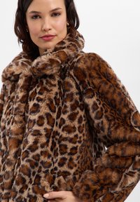 Płaszcz z futerka ekologicznego w panterkę, z kołnierzem zagiętym do środka, miękką w dotyku teksturą i luźnym krojem. Paleta kolorów obejmuje brązy i czernie.
