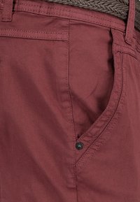 Burgundy cotton pants with a side pocket featuring a black button, detailed stitching, and a braided brown belt completing the look.