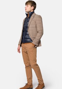 Marynarka w beżową kratę na granatowej kamizelce puchowej, zestawiona z khaki spodniami i brązowymi zamszowymi butami. Strukturalny, warstwowy wygląd.