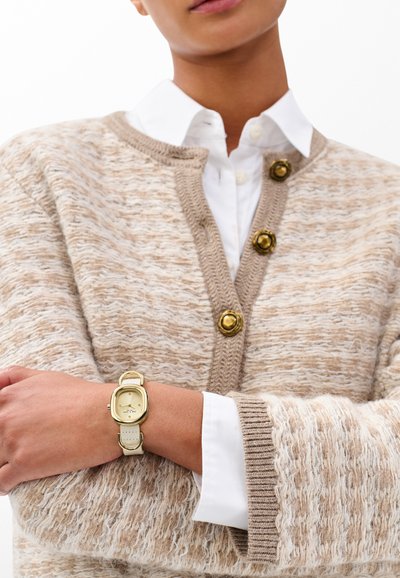 Beiger, strukturierter Cardigan mit goldenen Knöpfen über einem weißen Hemd. Am Handgelenk wird eine gold-weiße Uhr mit rechteckigem Zifferblatt getragen.
