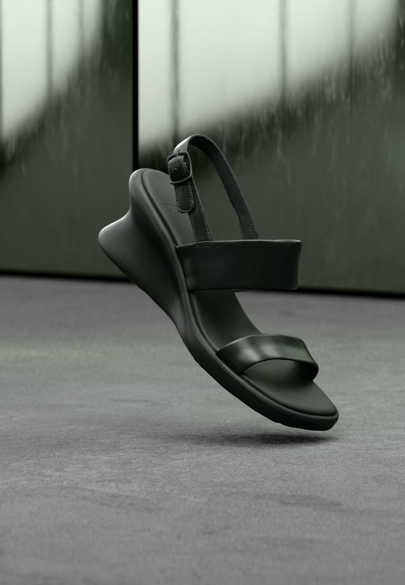 Sandale noire à bout ouvert avec deux brides et une boucle de cheville réglable, présentée flottant au-dessus d'une surface grise sur un fond vert flou.