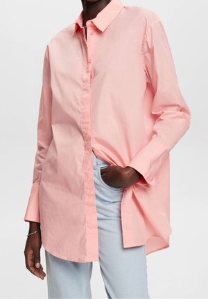 Donkere huidtint, gekleed in een losvallend, lichtroze overhemd met lange mouwen en lichtblauwe spijkerbroek, met één hand in de zak en een zilveren armband om de pols.