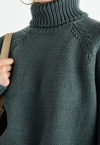 Pull tricoté vert foncé avec un col roulé côtelé haut, présentant un design texturé et des coutures d'épaule tombantes.
