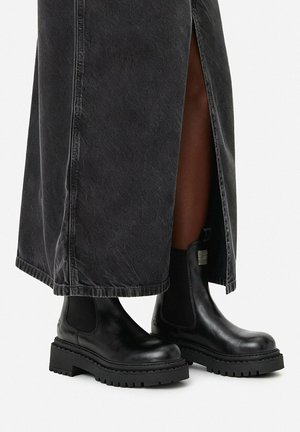Schwarze Chelsea-Stiefel aus Leder mit klobigen Sohlen, getragen zu einem langen schwarzen Rock mit seitlichem Schlitz, der nackte Beine zeigt.
