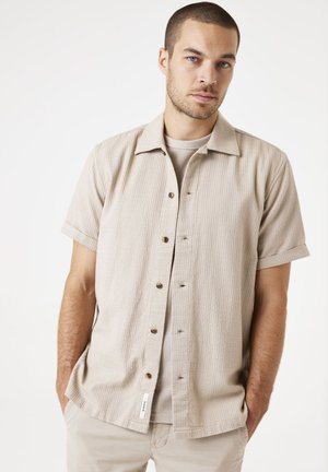 Chemise beige à manches courtes avec un motif texturé, bouton sur le devant et un col classique. Portée par-dessus une chemise de couleur claire. Étiquette visible au niveau de l'ourlet.