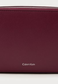 Portafoglio in pelle bordeaux con finitura texture, chiusura con zip e logo "Calvin Klein" in argento nell'angolo in basso a destra.